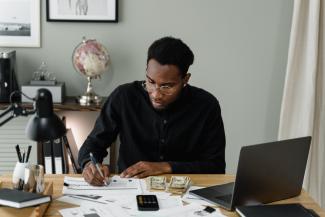 A young man sitting at a desk. There is a calculator and money on the desk, and the man is writing on a chart.